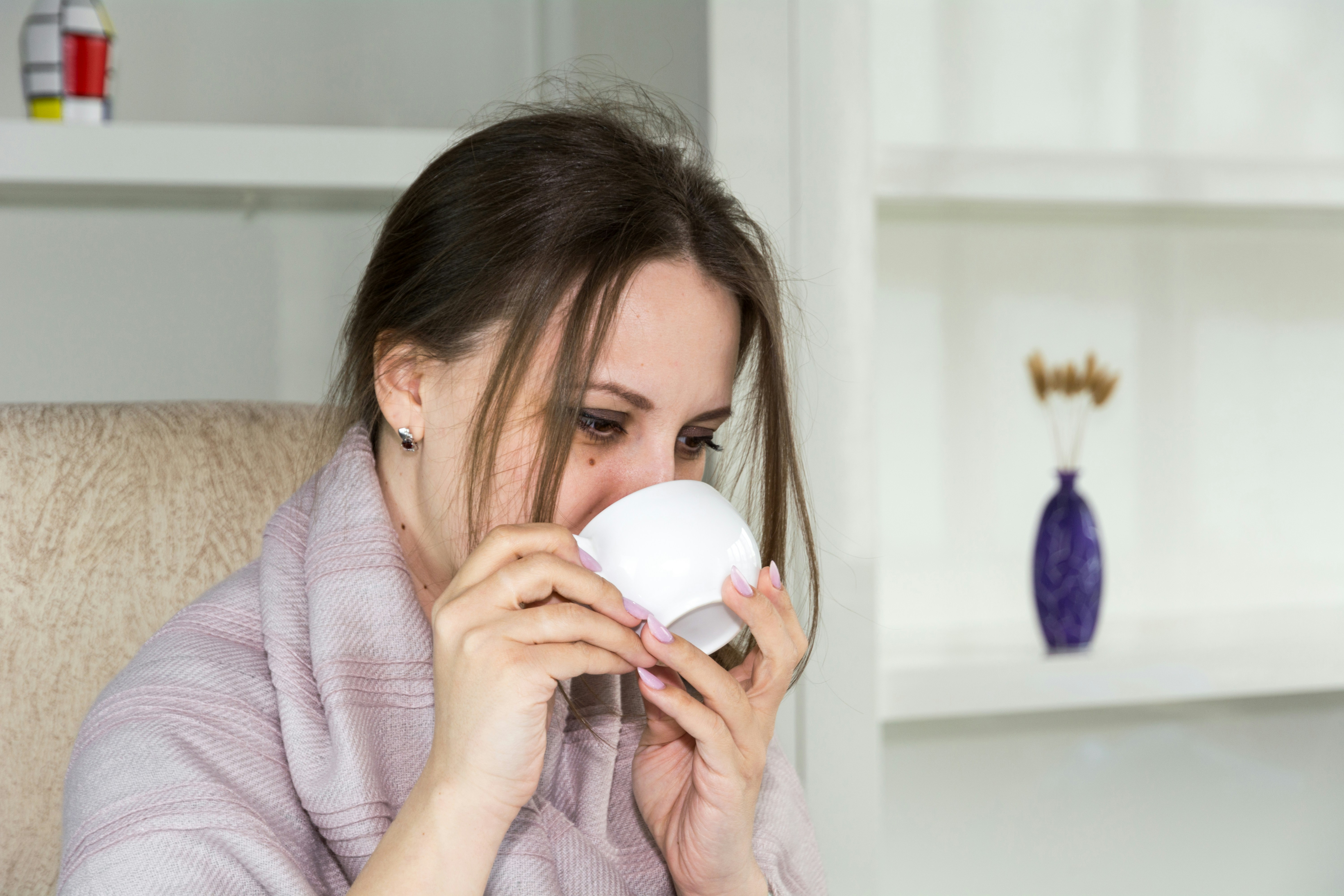A woman sitting quietly in a softly lit room, hands wrapped around a mug, looking out a window with a tired but determined expression.