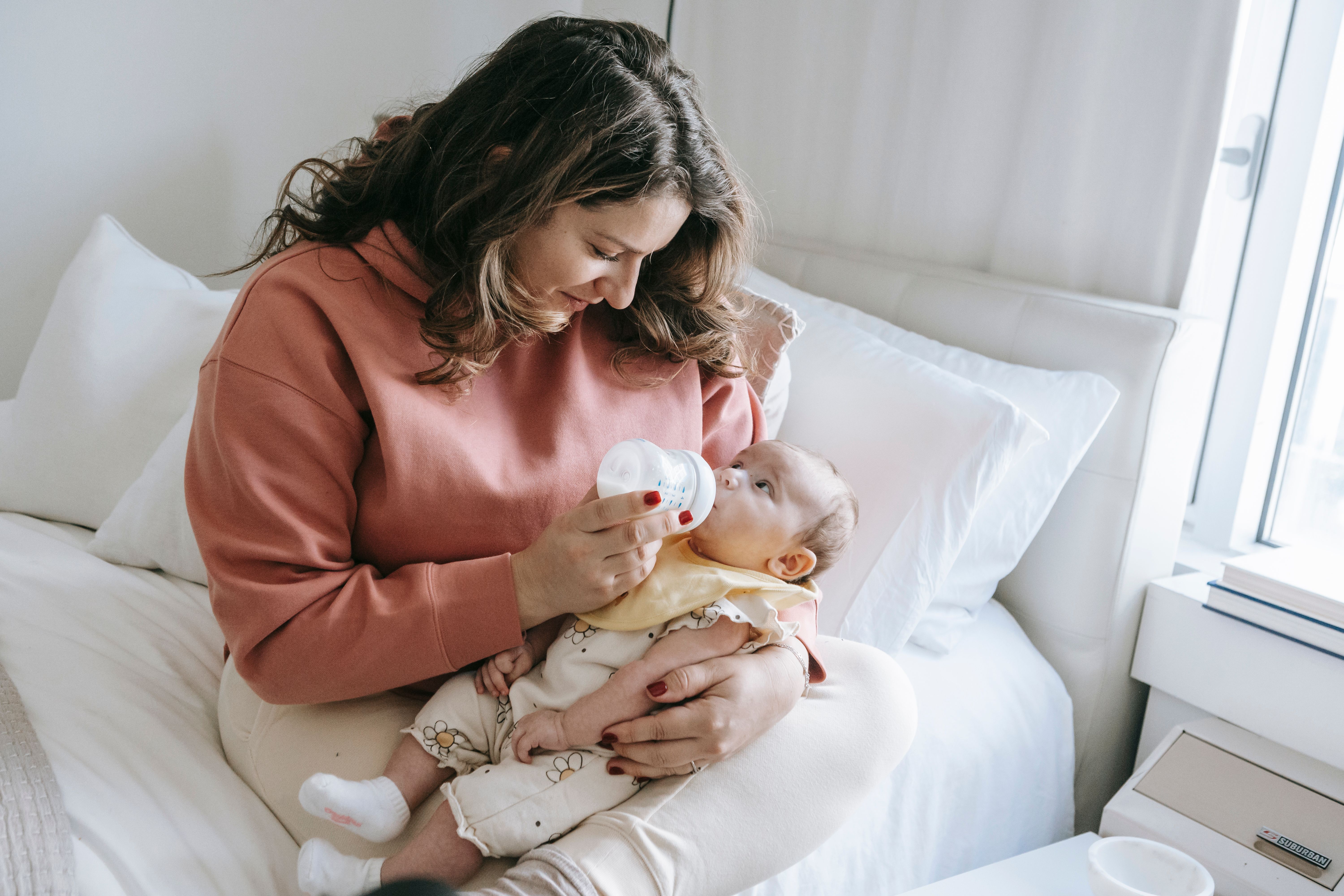 A parent gently holding a bottle while feeding their baby, looking down with a tender, thoughtful expression.